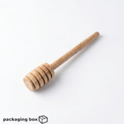 Hand holding a long handle wooden mixing stick dripping honey into a glass jar with cork lid, lavender flowers and bees in background