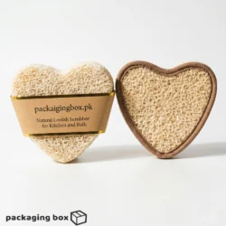 Two heart-shaped natural loofah scrubbers — one with kraft paper band label and one showing loofah texture with brown fabric border, for kitchen and bath use