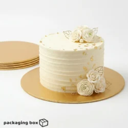 Golden Cake Board under a single-tier ivory buttercream cake with white roses and gold leaf decorations, with stacked gold cake boards visible in background by Packaging Box Pakistan
