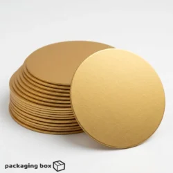 Golden Cake Board under a single-tier ivory buttercream cake with white roses and gold leaf decorations, with stacked gold cake boards visible in background by Packaging Box Pakistan