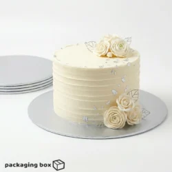 Silver Cake Board under a single-tier ivory buttercream cake with white roses and silver leaf decorations, with stacked silver cake boards visible in background by Packaging Box Pakistan
